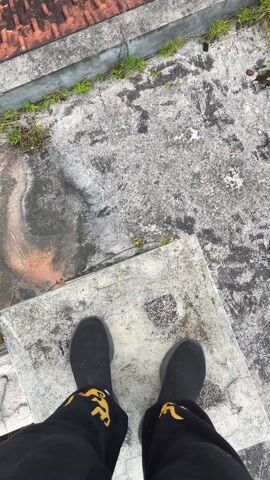 The video begins with a shot of a person's feet standing on a concrete surface with black shoes and patterned black pants. The camera pans upward to show a view of a building with a red-tiled roof, lush green vegetation, and a landscape of rolling hills. The sky is overcast with hints of sunlight breaking through the clouds. The setting appears to be a mountainous or hilly area with dense forests. The camera continues to pan upward, revealing a wider view of the landscape, showcasing the natural beauty of the environment.