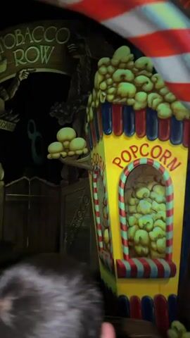 The video begins with a close-up of a popcorn cart, its bright yellow and red stripes and oversized popcorn decorations suggesting a carnival or amusement park setting. The camera then pans left, revealing a dark, dimly lit environment. A sign reading "TOBACCO ROW" with ornate lettering arches over the scene. Below the sign, a large wooden cutout depicts a figure holding several items, possibly cigars, with a box labeled "CIGARS" visible. To the left of the sign, and partially obscured by it, are three large, stylized yellow spheres, resembling oversized light bulbs or fruit.

As the camera continues to pan, the scene shifts to a stage-like area. Two anthropomorphic animal characters, one appearing to be a fox wearing a blue suit and hat, and another, possibly a cat, in a purple outfit, are positioned on a platform. Between them is a tall, brightly lit green structure with numerical markings from 0 to 50, resembling a measuring device or score board. The fox character is leaning on a cane, looking towards the scoreboard. The cat character is seated, looking forward. Several colorful pennants on poles are strung behind the characters, adding to the festive atmosphere. The background remains dark, with hints of wooden structures and more stylized elements. The overall impression is that of a themed attraction within a larger entertainment venue.