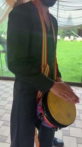 A man in a black robe and black pants plays a drum with a colorful strap. He is standing on a tiled floor. Behind him is a grassy area with a playground. The scene is outdoors, possibly under a tent.