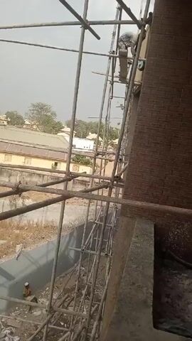 The camera is positioned on a building under construction. A section of scaffolding dominates the view, with a brick wall visible to the right. A worker is on the scaffolding at a higher level, appearing to be engaged in construction work. Another worker is sitting on the ground in the lower left corner. In the background, there are buildings and trees. The sky is overcast. The area beneath the scaffolding appears to be a construction site with debris.