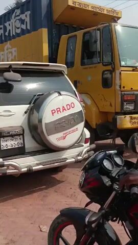 The video shows a man on a motorcycle waiting in traffic on a dirt road. The man is wearing a purple shirt, a lungi, and a head covering. He is facing away from the camera. In front of him is a white SUV with a spare tire on the back that says "PRADO". To his right is a large truck with a blue container on the back with yellow lettering on it. To his left is an auto rickshaw. The road is unpaved and dusty. Other vehicles and people are visible in the background.