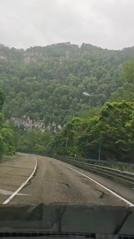 The video shows a car driving on a winding road through a mountainous region. The road is paved and has white lines marking the lanes. There are guardrails on the side of the road. The mountains are covered in lush green vegetation, and the sky is overcast. A bus passes in the opposite direction.