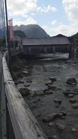 The video captures a river flowing through a village nestled in a mountainous region. On the left side of the frame, a wooden railing runs alongside a path. Beyond the path, people are visible walking and standing, and a few flags are planted. A covered wooden bridge spans the river in the middle ground. The river itself is shallow and rocky, with clear water flowing over stones. In the background, houses and trees are scattered on the slopes of the mountains, which are partly covered in clouds under a bright, cloudy sky. The camera pans slowly to the right, revealing more of the village and the natural landscape.