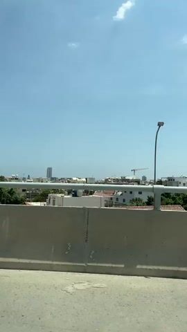 The video shows a view from a moving vehicle, looking over a concrete barrier towards a cityscape under a clear blue sky with scattered clouds. The cityscape consists of various buildings, including modern glass-fronted structures and more traditional residential buildings with terracotta roofs. Trees and a construction crane are also visible in the distance. The sun appears to be high in the sky, casting shadows and illuminating the scene. The overall impression is a daytime view of an urban or suburban environment.