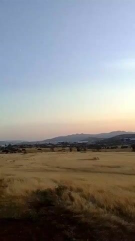 The video shows a wide shot of a field of dry grass with hills in the distance. The sky is clear and transitioning from blue to orange as the sun begins to set behind the hills. The sun's rays are visible, cutting across the sky. The camera is stationary.
