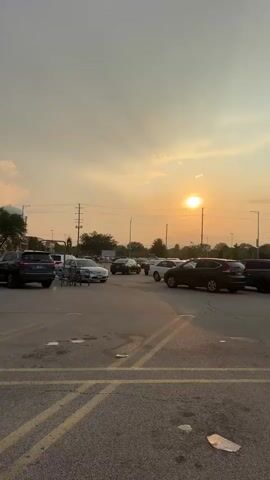 The video captures a parking lot scene at sunset. The sun, a bright orb, is visible in the sky, casting a warm, orange glow. The sky is partly cloudy, with some rays of sunlight piercing through. Several cars are parked and in motion within the lot. A shopping cart is also visible. In the background, there are trees and utility poles. The ground is paved asphalt with yellow parking lines. The overall impression is that of a common, everyday scene bathed in the beautiful light of a setting sun.