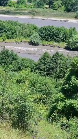 The video shows a wide shot of a river flowing through a lush green forested area. The river appears to be muddy and somewhat shallow, with visible banks and vegetation growing along its edges. In the distance, there are hills covered in trees, and some man-made structures can be seen on the far bank, possibly buildings or industrial facilities. The camera is positioned high up, looking down on the landscape, providing a panoramic view. There are no people or animals visible in the footage, and the focus is entirely on the natural environment and the river. The lighting suggests it is daytime, with sunlight illuminating the scene and casting shadows. The overall impression is one of a natural landscape with some signs of human presence in the background.