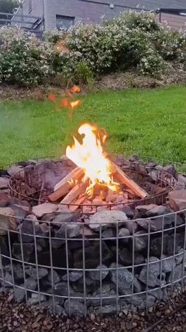 A close-up shot shows a fire burning in a metal cage filled with rocks. The fire is made of logs and flames are visible. A person's hand reaches into the frame from the right and adjusts one of the logs. The background shows green grass, bushes, and a building.