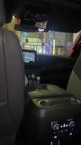 From the backseat of a car, the video shows a view of the vehicle’s dashboard and the illuminated street outside the front windshield at night. A navigation system is active on the dashboard screen. The exterior environment is wet, with raindrops visible on the windows. The camera then shifts its focus to the side window, revealing cars moving in the rain.