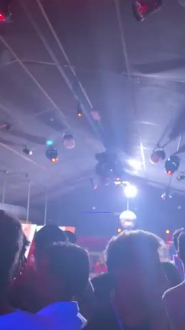 This video shows a crowded indoor scene, likely a nightclub or party, illuminated by alternating red and blue lights. Several disco balls are visible hanging from the ceiling, reflecting the lights. The camera focuses on the heads and shoulders of people dancing and socializing. Some individuals are looking towards the camera or at each other, while others are partially obscured by the crowd. The overall atmosphere appears lively and energetic, with the pulsating lights contributing to a dynamic ambiance. The video is short, with the lighting shifting from red to blue and back again.