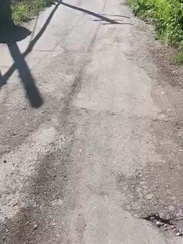 The video shows a dirt road in a rural area. The road is lined with fences and vegetation on both sides. In the distance, there are some buildings and hills. The sun is shining, casting long shadows across the road. The video appears to be taken from a moving vehicle, as indicated by the slight blur and the perspective. There are no people visible in the video.