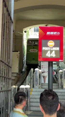 The video shows a green funicular train labeled "Dr.Max" ascending a steep track. To the right, a red digital sign displays "POSTI LIBERI 44," indicating 44 free seats. Two people are visible from the back, facing the train. The environment appears to be an outdoor station with a metal fence on the left and a covered entryway for the track. Green ivy covers the walls along the track.