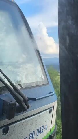 The video captures the front of a blue and white train locomotive in motion, seen from a slightly elevated, parallel viewpoint. The train's large windshield prominently reflects a bright blue sky dotted with large, cumulus clouds. Below the windshield, two black wipers and a blue horizontal grab handle are visible. A small white star sticker is affixed to the windshield, slightly off-center to the right.

The background showcases a picturesque rural landscape with expansive green fields and rolling mountains receding into the distance. A dark, textured side panel, likely from an adjacent train car or structure, partially frames the right side of the view. The weather is sunny and clear, suggesting daytime. The train is actively moving, accompanied by a continuous mechanical hum or whoosh in the audio.

Visible text on the lower front panel of the locomotive includes the partial number '80 042-0'. The setting is Karnare, Bulgaria, offering a serene view of the Bulgarian countryside from the perspective of a moving train.