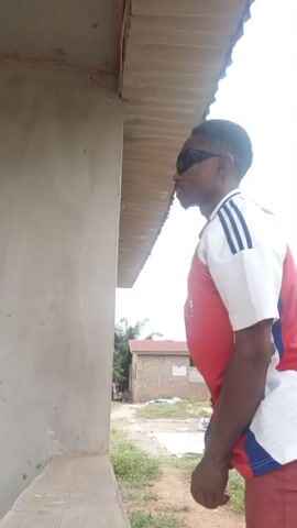 A person wearing a red and white soccer jersey, maroon pants, and sunglasses walks into view. He is standing next to a grey concrete building with a metal roof overhang. The person looks at the camera and points up in the air with one finger. There is a dirt yard with sparse vegetation and another building visible in the background. The sky is overcast.