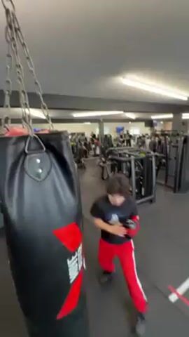 In a well-lit gym, a young man wearing a black t-shirt and red track pants is punching a black punching bag hanging from chains. He is wearing red boxing gloves and appears to be practicing a boxing sequence. The punching bag has a large red "X" and the word "HERO" printed on it. In the background, gym equipment and other people are visible, contributing to the bustling atmosphere of a gym. The floor is black with white line markings. The camera angle is low, looking up at the punching bag and the man.