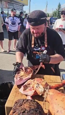 A man wearing a black t-shirt, a black cap, sunglasses, and a leather apron is carving a large piece of meat with a knife. He is also wearing black gloves and a headset. The meat is on a wooden cutting board, and there are other pieces of meat and a whole roasted pig on the board as well. In the background, there are several people standing around watching, and a building with a sign that says "MASS". The scene appears to be outdoors. The man is speaking into a microphone, and in the last few frames he asks, "What is the most delicious? The leg."