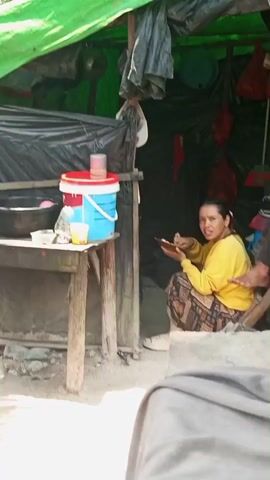 A woman in a yellow long-sleeved shirt and patterned pants is sitting on a low stool, eating from a plate held in her left hand. She is in a makeshift shelter with a green tarp roof and black plastic walls. A wooden table is to her left, with a blue and white bucket with a red lid, a smaller cup, and other items on it. The ground is covered in dirt and rocks.