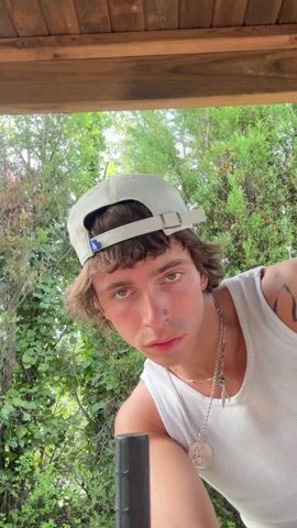 The video shows a young man standing under a wooden structure with dense green foliage in the background. He is wearing a white tank top, a backward white baseball cap, and several silver necklaces. He has light brown hair with bangs and green eyes. He is initially looking directly at the camera. He then straightens up and raises his right arm above his head with a clenched fist, striking a pose. A black cylindrical object is visible at the bottom left corner of the frame.