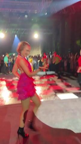 A group of people are dancing on a checkered floor in a dimly lit venue. The dance floor is illuminated by red and white lights. In the foreground, a woman in a pink, sequined dress is dancing. Behind her, another woman in a gold sequined dress is also dancing. Other people are visible in the background, some dancing and some watching. There are lights hanging from the ceiling and a stage with stairs on the left side of the frame. The overall atmosphere appears to be festive and energetic.