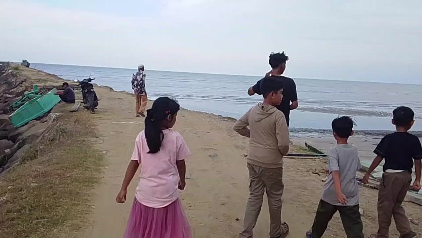 Several people are walking towards the ocean on a sandy path. In the foreground, a young girl in a pink shirt and tutu walks away from the camera. To her right are two boys and a taller person in a beige shirt and pants. In front of them is a person in a black shirt. To the left, a person wearing a kufi and patterned shirt is walking the same direction. Farther to the left is a motorcycle parked next to a green dinghy and a blue fishing boat. Two people are sitting on the side. The sky is overcast, and the water is calm.