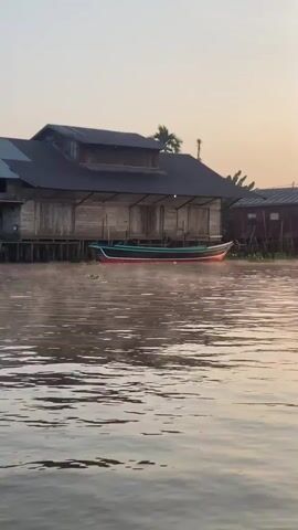 The video shows a body of water with a stilt house on the left, a boat tied to the house, and other stilt houses in the background. The sky is hazy with a warm, orange hue, suggesting sunrise or sunset. The water is calm with ripples, and there is some vegetation floating on it. The stilt houses are made of wood and appear weathered. The boat is long and narrow, painted in red, green, and black. The overall atmosphere is peaceful and serene.
