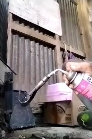 A person's hand holds a pink spray can with a black attachment, directing a flame towards a black object. The background features a corrugated metal wall and some boxes.