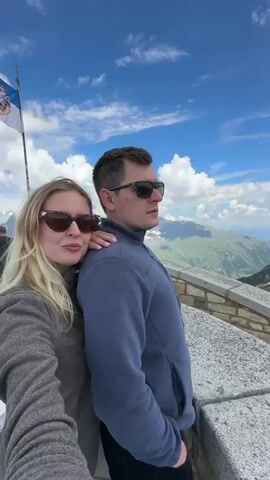 A man and a woman are standing close together, with the woman leaning her head on the man's shoulder. They are both wearing sunglasses and looking out at a mountainous landscape. The sky is blue with white clouds. There is a flag pole to the left of the frame. The man is wearing a blue sweatshirt and the woman is wearing a grey coat.