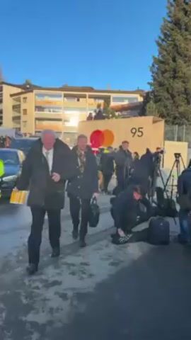 The video shows two men walking towards the camera on a wet street. The man in front wears a black coat and the man behind wears a dark coat and a plaid scarf. Behind them, a group of people stand near a large structure with the number 95 and a logo. A worker in a bright yellow safety vest stands by the street, signaling for a black car to move forward. The background features buildings and a clear blue sky. Numerous people are visible in the background, moving and standing in groups. Several cameras and equipment are set up. The overall scene suggests a public event or gathering.
