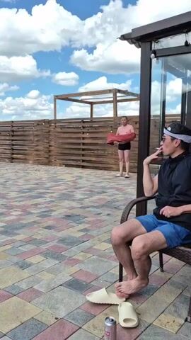 A man in a black vest and blue shorts sits in a chair, smoking and wearing a headband and sunglasses. His feet are bare, and he wears white slides. In the background, another person is seen standing near a wooden fence with a red towel. The scene is an outdoor patio with patterned paving stones and a wooden fence. The sky is blue with white clouds. A second clip shows the same patio and fence, with people walking near a body of water.