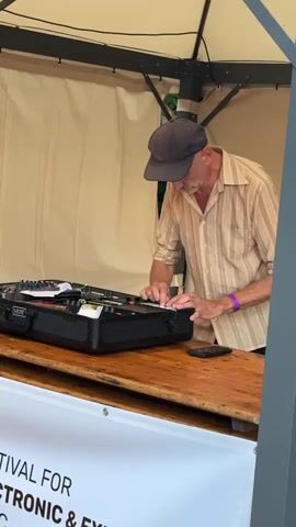 A man wearing a cap and a patterned shirt is standing behind a wooden table inside a tent. He is operating some electronic music equipment, which is housed in a black case. A banner with text on it, possibly indicating an event, is displayed on the front of the table. There are speakers and other equipment visible around the tent, suggesting an outdoor music event. The man is focused on the equipment, with his hands on the controls. The background shows trees and greenery.