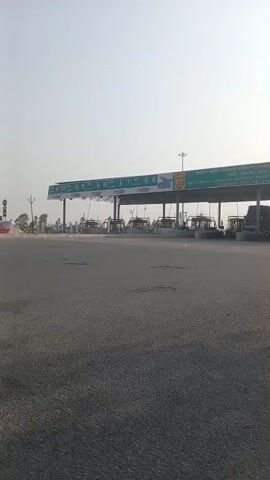 The video begins with a static shot of a tollbooth on National Highway 27. The sky is clear and light. A motorcycle with two riders passes in front of the tollbooth. The camera then pans to the right, showing a road leading to a group of large dump trucks parked near a small white building. A motorcycle also passes by this area.
