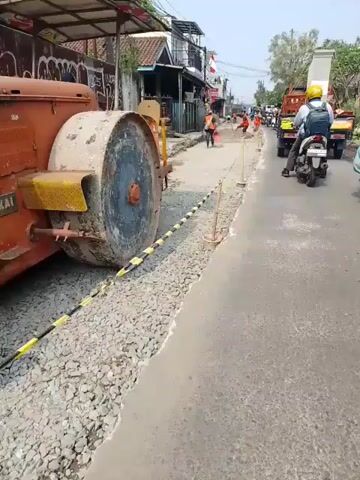 The video captures a street construction scene. In the foreground, a large, orange steamroller is parked on the left side of the road, its heavy roller partially obscuring the view. Yellow and black caution tape is strung along a line of gravel, marking off the construction area. The road itself appears to be undergoing resurfacing, with patches of exposed gravel and some areas of compacted earth. Several construction workers in orange vests are visible further down the road, some appearing to be shoveling or leveling the ground. On the right side of the road, a motorbike with a rider wearing a blue shirt and a yellow helmet is moving away from the camera, followed by another motorbike. A white truck with an open bed is also present further up the road. The background shows a row of buildings, some with balconies and awnings, and overhead electrical wires. The sky is clear and bright, suggesting it is daytime. The overall impression is of an active, albeit somewhat congested, road repair operation.