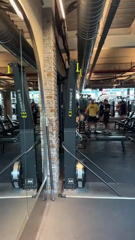 The video shows the interior of a gym in Şişli, Turkey. Two rowing machines, branded "Concept2," are prominently featured in the foreground. They are positioned next to a brick wall and a large mirror, which reflects the gym environment. In the background, several people are visible, some engaging in workouts and others standing around. The lighting suggests it is daytime. The overall atmosphere appears to be active and typical of a gym setting. Text visible on the rowing machines includes "Concept2" and what appears to be a model number "44" on one of them.