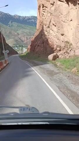 The video shows a view from inside a vehicle looking out at a paved road winding through a narrow canyon. The canyon walls are steep and made of reddish-brown rock. Green vegetation grows on the slopes and at the bottom of the canyon. In the distance, mountains are visible, and a small village or town can be seen nestled in a valley. The sky is blue with some clouds. The road has a white line marking the center and a white line on the right edge. Streetlights are present along the road. The front windshield of the vehicle is visible at the bottom of the frame, along with what appear to be windshield wipers. The camera is moving forward, indicating the vehicle is in motion.