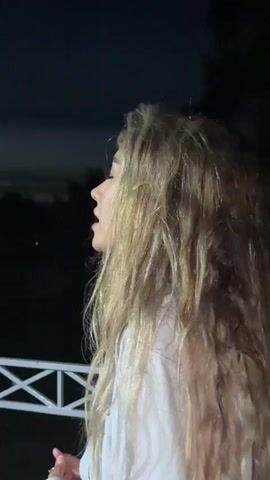 The video shows a young woman with long, blonde, wavy hair in profile against a dark background. She is wearing a white shirt. She appears to be singing or speaking with her mouth open. A white railing is visible in the lower left corner of the frame. The lighting suggests it is nighttime or a very dark indoor setting.
