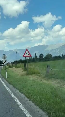 This is a vertical, wide-angle shot taken from a vehicle traveling on a road in Mitterhofen, Austria. The scene depicts a rural landscape with mountains in the background. The road has a white line marking the lane.

To the right of the road, there is a grassy field with some dry patches and wooden fence posts. A sign with a red triangle and a deer silhouette indicates a deer crossing zone for the next 1.7 kilometers. Further down the road, there are other signs, including one with a black and white circle and diagonal line, which typically signifies a speed limit restriction or the end of a restriction. Another circular sign above it displays the number "57.6."

In the distance, a range of majestic mountains, some with snow-capped peaks, dominates the horizon. Scattered evergreen trees are visible on the lower slopes of the mountains. The sky is a vibrant blue with fluffy white cumulus clouds, suggesting a pleasant day, likely in the late morning or early afternoon. A few cars and a bus are visible on the road ahead, indicating moderate traffic. The overall mood is one of travel through a scenic, mountainous region.