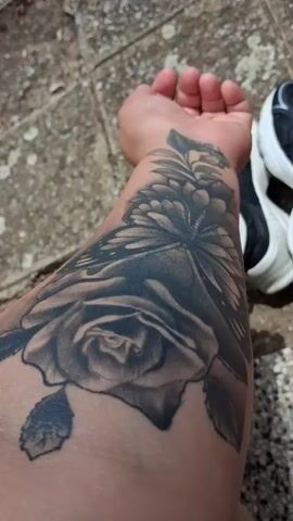 The video shows a close-up view of a tattoo on a person's forearm. The tattoo features a large rose in black and gray shading, with a butterfly positioned above it, also in black and gray. There are also some leaves and possibly flower buds depicted. The person's arm is resting on a textured surface that appears to be cobblestones or a similar outdoor ground material. In the background, a pair of black and white sneakers are visible, with white socks peeking out. The lighting suggests it is daytime, possibly outdoors. The camera movement is minimal, mostly static with slight adjustments in angle and focus.