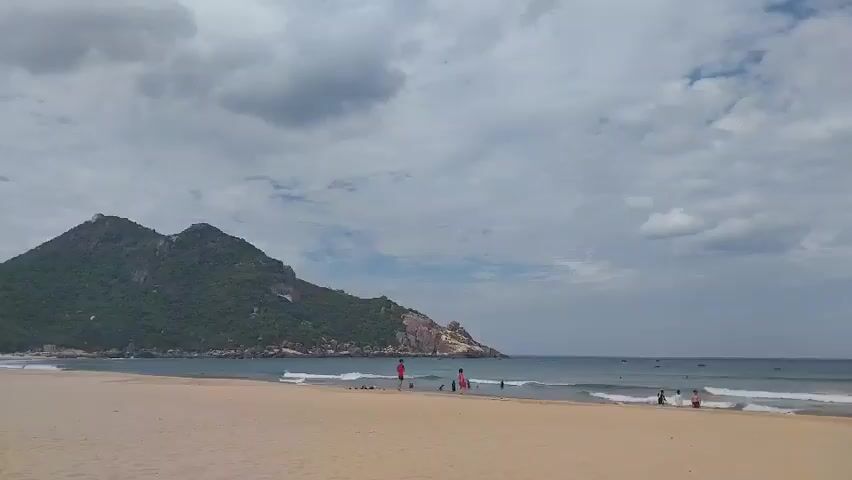 A wide shot of a sandy beach shows a calm ocean with gentle waves. In the distance, a lush green mountain rises from the coastline. The sky is partly cloudy with grey and white clouds. Several people are scattered on the beach and in the shallow water. On the left side of the beach, a person in a red shirt stands near the water's edge, while a person in an orange dress is further down the beach. In the middle ground, a few individuals are wading in the ocean. On the right side of the frame, a small group of people are gathered near the water. A few small boats are visible on the horizon. The sand is light brown and appears smooth. The overall scene is peaceful and suggests a leisurely day at the beach.