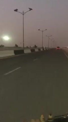 A first-person perspective video shot from a moving vehicle, presumably a motorcycle, shows an asphalt road stretching forward under a dusky sky. The road is flanked by concrete barriers, with evenly spaced streetlights on either side.  A vehicle, likely a truck, is visible in the distance with its tail lights illuminated. Along the barriers, dark silhouettes of people can be discerned. The motorcycle's handlebar is visible in the lower portion of the frame, suggesting the rider's viewpoint.