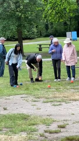 A group of people are playing bocce ball in a park. There are at least six people visible. In the foreground, a woman with long dark hair is bending down to roll a bocce ball. She is wearing a light blue jacket and dark pants. To her left, there are three blue bocce balls on the grass. A man in a black jacket and khaki pants is standing behind her, holding a yellow bocce ball. To the right of the woman, an older woman in a blue jacket and dark pants is bending down, and another older woman in a pink jacket and a hat is standing. Two older men are visible in the background, one in a blue jacket and jeans, and another in a dark jacket and cap. The game is taking place on a grassy area with some gravel in the foreground. Trees and foliage are visible in the background, along with two blue structures that appear to be portable restrooms or storage units. The weather appears overcast.