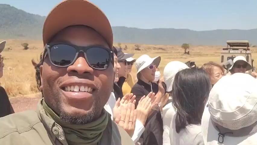 A man with sunglasses and a tan baseball cap is taking a selfie-style video with a group of people behind him. Most of them are wearing hats. They are clapping and cheering. A safari vehicle is parked behind them. The landscape appears to be a savanna, with dry grass and scattered trees. Mountains are visible in the distance.