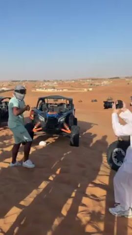 The video shows two people in a desert landscape taking photos. One man is wearing a light green outfit, white sneakers, and a black and white patterned headscarf. He dances briefly and poses for photos. Another man is wearing traditional Arab clothing, including a white robe and a similar headscarf. He is holding a professional camera and taking pictures. In the background, there are off-road vehicles parked on the sand. The sky is clear and blue, suggesting it's daytime. In the distance, there is a view of a town. The overall setting seems to be a tourist attraction or a staged photo shoot in a desert environment.
