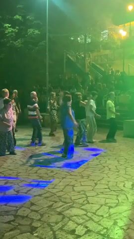 A group of people are dancing in an outdoor area at night. They are illuminated by colored lights projected onto the stone-paved ground. In the background, more people are gathered, and there are trees and a staircase visible. Some of the dancers are in the foreground, while others are further back. The atmosphere appears festive.