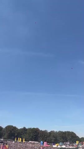 The video opens with a wide shot of a clear blue sky with a few small red objects visible, possibly kites or lanterns. The camera then pans down to reveal a large crowd of people gathered outdoors, likely at a festival or event. In the distance, there are stages and tents, along with trees bordering the area. The crowd is dense, with many individuals wearing colorful clothing and hats. A flag on a pole is prominently displayed in the foreground, featuring text and an image. The overall atmosphere appears to be festive and sunny.
