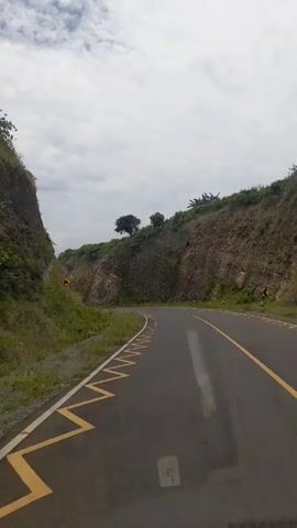 The video shows a car driving along a winding road through a hilly landscape. The road is paved with asphalt and has yellow lines on both sides. There is grass and shrubs on either side of the road, and the hills are covered in vegetation. The sky is cloudy. The audio contains speech, but the content is unknown.