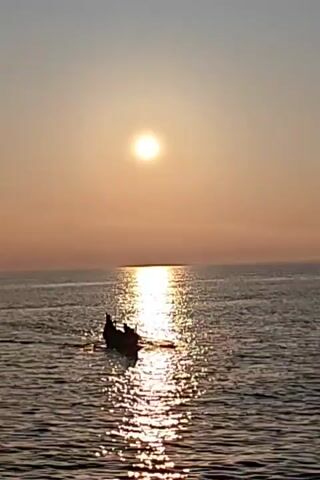 A distant boat, silhouetted against the bright sun and its reflection on the water, moves from right to left across the frame. Several figures are visible in the boat. The water is calm, with gentle ripples reflecting the sunlight. The sky is a gradient of light blue to orange, indicating either sunrise or sunset.