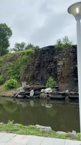 The video shows a rocky cliff face with greenery growing on and around it. A body of water is visible at the base of the cliff, reflecting the scene above. A modern streetlamp with a light on is positioned in the foreground to the right of the frame. The sky is overcast with clouds. The water appears to be trickling down sections of the rock face.