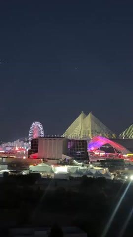 This video captures a night scene featuring a large Ferris wheel, a brightly lit bridge, and several buildings or structures with colorful illumination. Tents and smaller buildings are visible in the foreground. The sky is dark, indicating nighttime, and a full moon is present. The overall impression is of a lively event or festival taking place in an urban or entertainment complex.