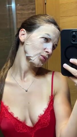This is a close-up selfie of a young woman with a sheet mask on her face, taken in what appears to be a bathroom. She is wearing a red lace-trimmed camisole and a silver necklace with a small pendant. She is holding a black smartphone in her right hand, angled to capture her reflection in a mirror. The background shows a wooden textured surface, likely part of a cabinet or wall. The lighting suggests it could be daytime, possibly in the morning or afternoon. The woman's expression is neutral, and the focus is on her and the sheet mask, indicating a personal moment of skincare. The overall impression is one of self-care and relaxation.