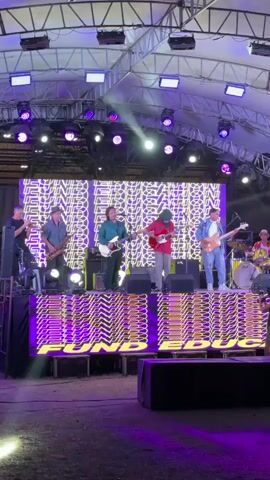 A band is performing on a stage outdoors. The stage is covered by a white tent-like structure with lights. In the background, a large LED screen displays abstract purple and white patterns. Below the screen, a yellow box with purple, wavy text reads "END SYSTEM STAGNA". Six musicians are on stage. From left to right, a person in a black shirt and hat plays a trumpet. Next, a person in a blue shirt and hat plays a saxophone. In the center, a person in a teal shirt plays a white electric guitar. To their right, a person in a red shirt and hat plays a red electric guitar. Further right, a person in a blue shirt and hat plays a brown electric guitar. On the far right, a person in a grey shirt plays a bass guitar. Behind the bass player, a drummer can be seen. Several speakers are placed on the ground in front of the stage. The ground in front of the stage appears to be dirt or gravel. Lights from the stage are illuminating the ground. The overall atmosphere appears to be a live music event.